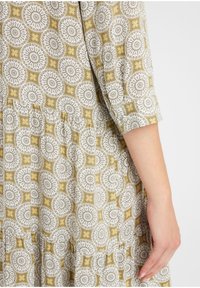 Rochie cu un model floral în nuanțe de galben și alb, cu mâneci scurte și un tiv volanat; materialul pare ușor, cu o suprafață texturată.
