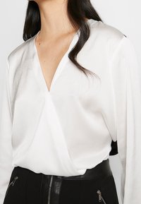 Woman wearing a white long-sleeve V-neck blouse tucked into high-waisted black pants with front zipper and zipper pockets.