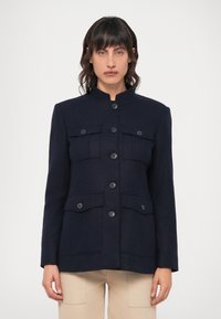 Tengerészkék gyapjú kabát magas gallérral, hat elülső gombbal és két zsebbel a mellkas tájékán. Jellemzi a testreszabott szabás és a sima textúra.