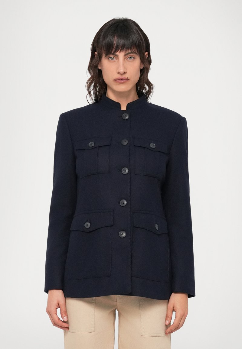 Tengerészkék gyapjú kabát magas gallérral, hat elülső gombbal és két zsebbel a mellkas tájékán. Jellemzi a testreszabott szabás és a sima textúra.