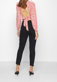 Red floral blouse with long sleeves, open back tied with bows, paired with high-waisted black jeans featuring a cropped, frayed hem. Black heels.