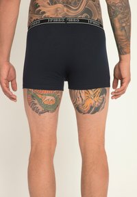 Boxer-briefs bleu marine avec une taille noire arborant le logo "JP1880". Les jambes découvertes présentent des tatouages colorés détaillés d'un tigre et d'un dragon.