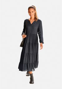 Schwarzes Maxikleid mit langen Ärmeln, V-Ausschnitt und gestuftem Rüschensaum. Besitzt ein zartes Muster und wird mit schwarzen Schnürstiefeln und einer kleinen Tasche kombiniert.