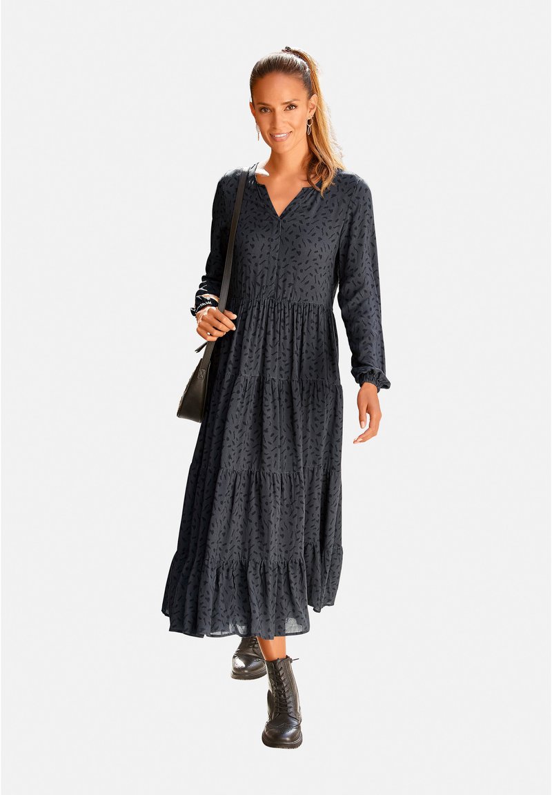 Schwarzes Maxikleid mit langen Ärmeln, V-Ausschnitt und gestuftem Rüschensaum. Besitzt ein zartes Muster und wird mit schwarzen Schnürstiefeln und einer kleinen Tasche kombiniert.