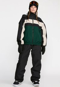 Waterproof snowboard jacket in dark green, beige, and black, with zip closure, adjustable cuffs, and a relaxed fit. Paired with black cargo pants.