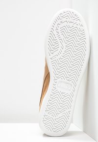 Tenisky s vrchní částí z béžové semišové kůže, bílé gumové podešve s klikatým vzorem. Mezipodešev je hladká a zakřivená, obsahuje detail s logem.