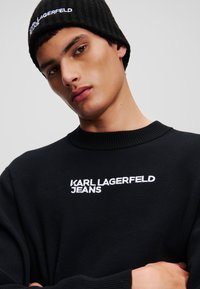 Fekete bordás sapka fehér hímzett "KARL LAGERFELD JEANS" logóval. Fekete kereknyakú pulóver hasonló hímzéssel a mellrészen.
