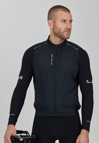 Schwarze Radweste mit hohem Kragen, Frontreißverschluss und reflektierenden Akzenten. Getragen über einem langärmeligen schwarzen Shirt, mit einem Fahrradhelm in der Hand.