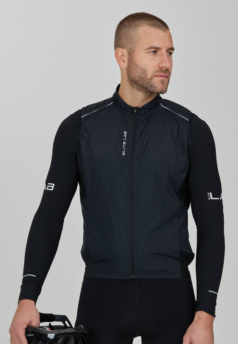 Schwarze Radweste mit hohem Kragen, Frontreißverschluss und reflektierenden Akzenten. Getragen über einem langärmeligen schwarzen Shirt, mit einem Fahrradhelm in der Hand.