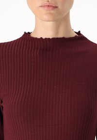 Bordo rebrasto majica od mekanog materijala, s visokim izrezom sa valovitim rubom. Tekstura je glatka s vertikalnim prugama.