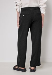 Zwarte, wijde broek met een gestructureerde stof en elastische taille. Heeft twee achterzakken en een klein merklabel nabij de tailleband.