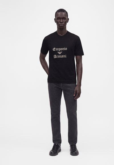Άνδρας φοράει μαύρο t-shirt Emporio Armani, σκούρο γκρι τζιν και μαύρα αθλητικά παπούτσια, στέκεται μπροστά σε απλό φωτεινό φόντο.