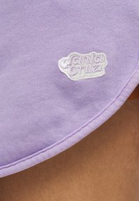 Ύφασμα σε χρώμα λεβάντας με λευκή κεντημένη ετικέτα λογότυπου "Santa Cruz" και καμπύλο ραμμένο στρίφωμα πάνω από γυμνό δέρμα.