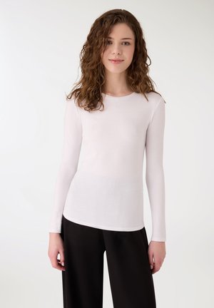 Giovane donna con capelli ricci castani, che indossa un top bianco a maniche lunghe e pantaloni neri, in piedi davanti a uno sfondo bianco uniforme.