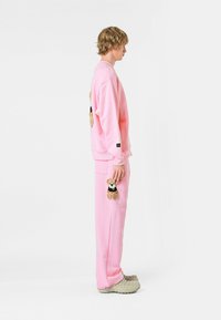 Sudadera y pantalones rosas hechos de un tejido suave, con aplicaciones de osito de peluche en el pecho y en el bolsillo. Llevado con zuecos grises texturizados.