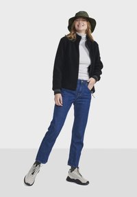 Crna flis jakna, bijeli turtleneck, plave traperice, zelena kacepe kapica i sivi tenisice s crnim potplatima, prikazani u punom tijelu.