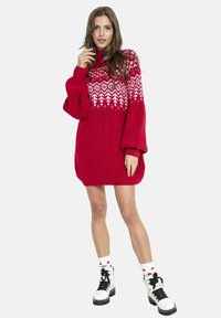 Czerwona oversize sukienka swetrowa z białymi geometrycznymi wzorami i wysokim kołnierzem, zestawiona z białymi botkami na sznurowadła i skarpetkami w gwiazdy.