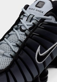 Κοντινή λήψη μαύρου και γκρι αθλητικού παπουτσιού Nike Shox που επικεντρώνεται στα κορδόνια, το πλεκτό ύφασμα και το ραμμένο λογότυπο Nike στο πλάι.