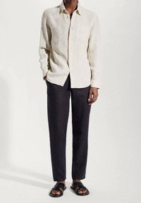 Persona che indossa una camicia a maniche lunghe beige con bottoni, pantaloni neri e sandali neri aperti, su uno sfondo chiaro e semplice.