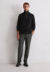 Pull à col zippé noir, pantalon à motifs gris et chaussures noires. Le matériau semble lisse, avec des poignets et une taille côtelés.