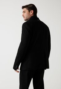 Zwarte colbert, slim fit, gladde textuur, enkele split, mouwen met drie knopen, minimale branding op de achterkant, weergegeven vanuit een iets gedraaide hoek.