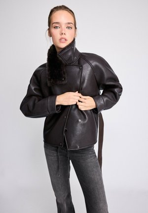 Jeune femme fermant une veste en cuir noire à col montant avec détail en fourrure, portant un jean gris foncé sur un fond clair uni.