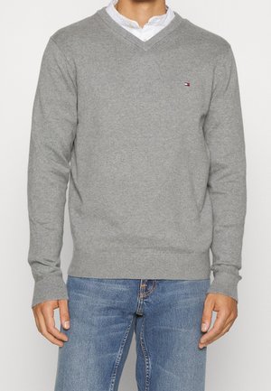 Maglione grigio a V in tessuto lavorato a maglia con polsini e orlo a costine, dotato di un piccolo logo sul lato sinistro del petto. Indossato sopra una camicia bianca.