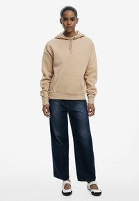Felpa beige con tasca frontale e coulisse, abbinata a jeans scuri larghi e scarpe slip-on bianche. Design semplice e informale.