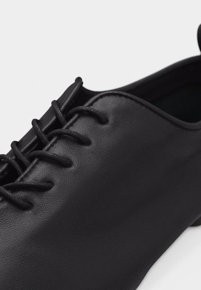 Gros plan sur une chaussure à lacets en cuir noir, à finition mate et lisse, avec un bout arrondi.