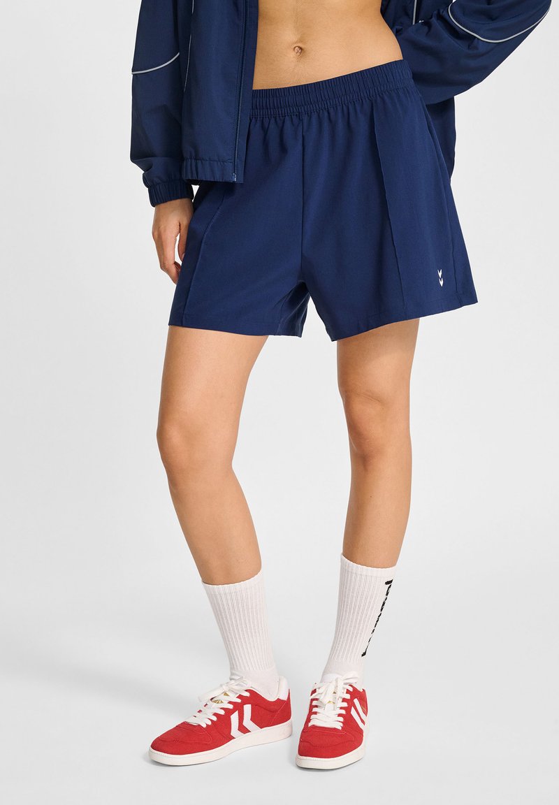 Pantaloni sportivi blu navy con cintura elastica e pieghe laterali. Indossati con scarpe da ginnastica rosse e calzini bianchi a manica corta con dettagli neri.