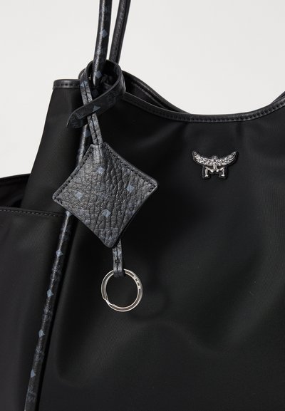 Saco tote preto com etiqueta de couro texturizado e anel prateado, apresentando um pequeno emblema prateado com asas perto da borda superior.