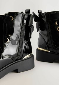 Botines de charol negro con un acabado brillante, acentos de hardware dorado, cremallera frontal y detalle decorativo de lazo negro en los laterales.