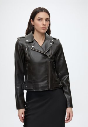 Femme aux cheveux foncés portant une veste de motard en cuir noir ajustée avec des zips et boucles argentés, debout devant un fond clair uni.