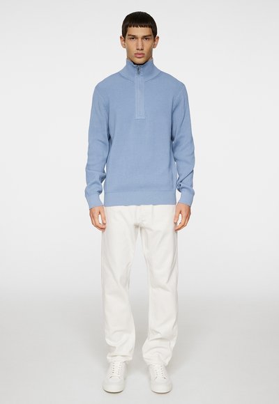 Hellblaues Reißverschluss-Sweatshirt mit gerippter Textur, hohem Kragen und langen Ärmeln, kombiniert mit weißen Hosen und weißen Sneakern.