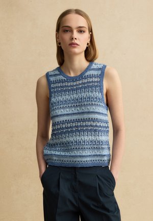 Vrouw met stijl blond haar draagt een blauw patroon mouwloos gebreid topje en zwarte broek, staand tegen een beige achtergrond.