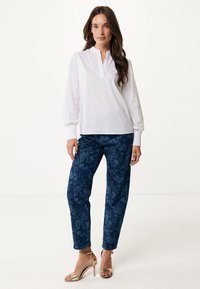 Blusa bianca a maniche lunghe con colletto a placca, abbinata a jeans blu navy con stampa floreale e sandali con tacco dorati. Tessuti morbidi, vestibilità rilassata.