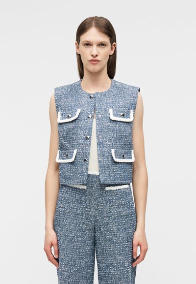 Γυναίκα που φοράει μπλε γιλέκο από τουίντ χωρίς μανίκια με τσέπες με καπάκια και κουμπιά, ταιριαστό παντελόνι υψηλής μέσης και λευκό τοπ από μέσα.