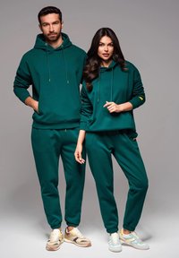 Felpe e pantaloni in cotone verde indossati da due modelli. Le felpe presentano cordini, mentre i pantaloni hanno polsini elasticizzati. Entrambi hanno dettagli gialli.
