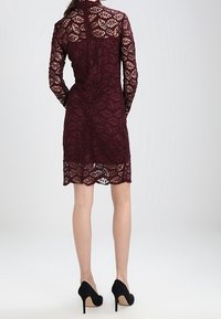 Burgundské krajkové šaty s dlouhými rukávy, květinovým vzorem a délkou do poloviny stehna; doplněné černými lodičkami na vysokém podpatku.