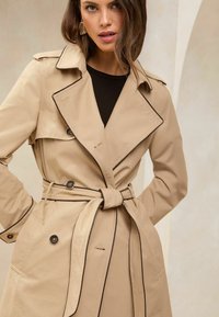 Bezs színű, strukturált fazonú trench coat, övvel a derekán. Kontrasztos fekete díszítésekkel és nagy gombokkal. Sima textúra, térdhosszúságú.