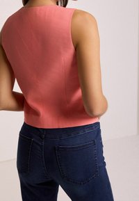 Top senza maniche color corallo con una texture liscia, scollatura alta e orlo dritto, abbinato a jeans in denim blu scuro con tasche posteriori.