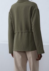 Olive green, long-sleeve jacket with a textured finish and a slightly loose fit. Features a collar and a back waistband for shaping.