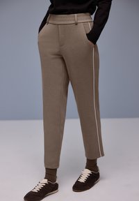 Beige broek met een ruime pasvorm, voorzien van een witte zijstreep, elastische tailleband en zijzakken, gecombineerd met donkerbruine schoenen.