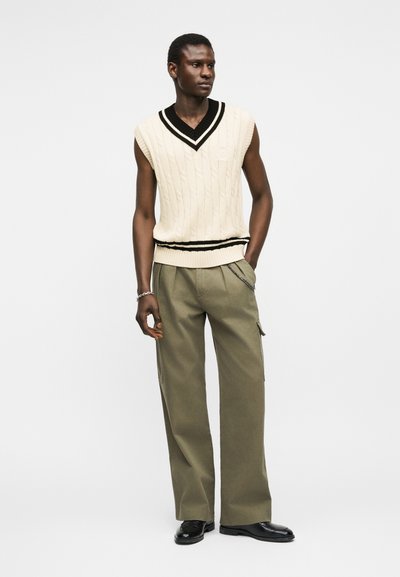 Άνδρας που φοράει λευκό μαν sleeveless πουλόβερ με πλεκτό σχέδιο καλωδίου και μαύρο τελείωμα, ελαιοπράσινο παντελόνι με φαρδιά πόδια και αλυσίδα στην τσέπη, και μαύρα παπούτσια, στέκεται με τα χέρια στις τσέπες.
