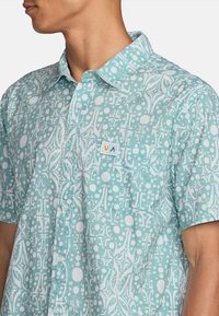 Light blue short-sleeve shirt featuring a white abstract pattern, button-up front, and a small logo patch on the chest. Lightweight fabric.