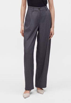 Femme portant un pantalon large taille haute gris foncé avec des plis à l'avant et des chaussures beiges à bout pointu, debout devant un fond uni.
