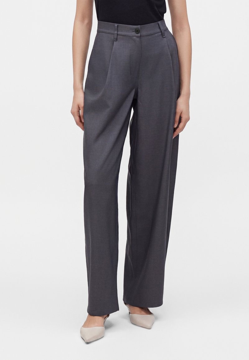 Femme portant un pantalon large taille haute gris foncé avec des plis à l'avant et des chaussures beiges à bout pointu, debout devant un fond uni.