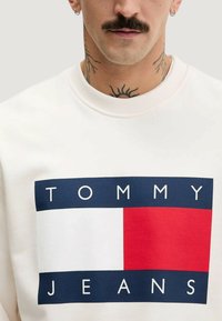 Vit sweatshirt med en marinblå, röd och vit rektangulär logotyp med texten "TOMMY JEANS" i fetstil, ribbad rundhalsad avslutning.