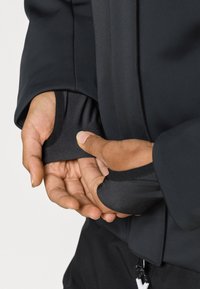 Čierna elastická bunda s rebrovanými manžetami a hladkou tkaninou. Detail rúk upravujúcich manžetu, ktorý zdôrazňuje funkčné dizajnové detaily.