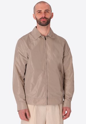 Uomo con barba corta che indossa una giacca marrone chiaro con zip e pantaloni beige, in piedi davanti a uno sfondo chiaro uniforme.